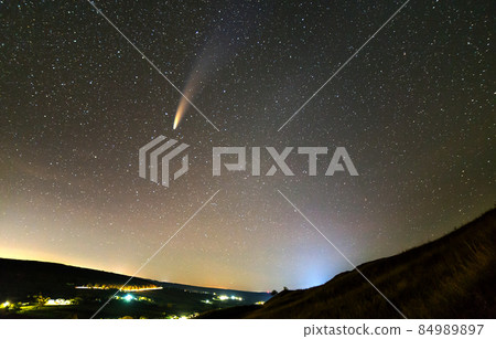 Summer night in mountains with starry dark blue cloudy sky and C/2020 F3 (NEOWISE) comet with light tail and bright lights of village. 84989897