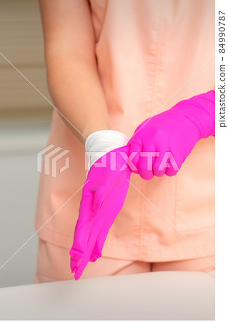 Hand of beautician puts on sterile pink gloves prepares to receive clients indoors. Hand of beautician puts on sterile pink gloves prepares to receive clients indoors. 84990787
