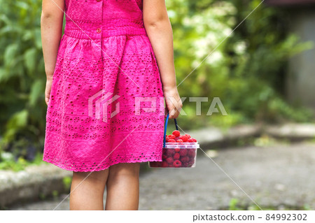 Little girl in dress child hand holdind small basket of ripe raspberries. Close-up 84992302