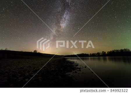 Landscape of pebbles river bank, trees on horizon, bright stars and Milky Way galaxy in dark sky reflected in quiet water. Beauty of nature concept. 84992322