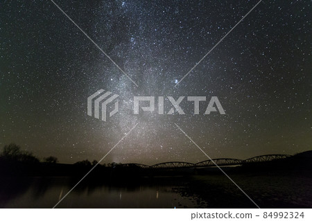 Landscape of pebbles river bank, trees on horizon, bright stars and Milky Way galaxy in dark sky reflected in quiet water. Beauty of nature concept. 84992324