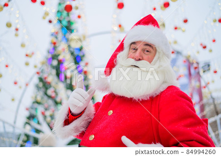 Portrait of an elderly man dressed as santa claus on the background of a christmas tree outdoors. 84994260