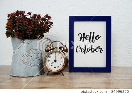 Hello October typography text with alarm clock and artificial plant decoration on wooden table and white wall background 84996260