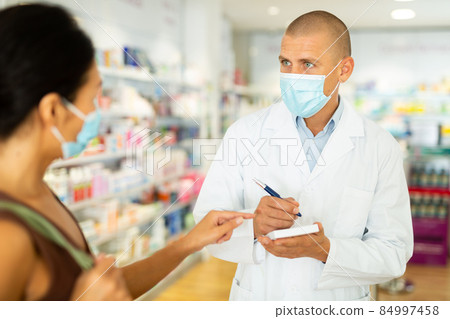 Pharmacist in protective mask writes in notebook the name of the drugs that buyer tells him to 84997458