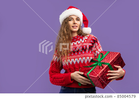 Beautiful smiling girl wears Santa hat holds red gift box on purple background. Christmas party Beautiful smiling girl wears Santa hat holds red gift box on purple background. Christmas party 85004579