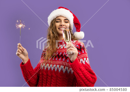 Happy girl wears Santa's hat drinks champagne holds sparkler on purple background. Christmas party Happy girl wears Santa's hat drinks champagne holds sparkler on purple background. Christmas party 85004580