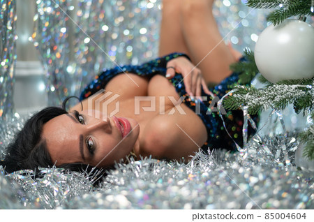 Sexy brunette in a black sequin dress by the Christmas tree. A woman with a large seductive breast lies among the silvery sequins Sexy brunette in a black sequin dress by the Christmas tree. A woman with a large seductive breast lies among the silvery sequins 85004604