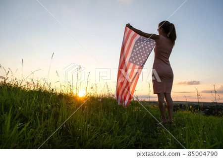 Happy young woman posing with USA national flag standing outdoors at sunset. Positive female celebrating United States independence day. International day of democracy concept. Happy young woman posing with USA national flag standing outdoors at sunset. Positive female celebrating United States independence day. International day of democracy concept. 85004790