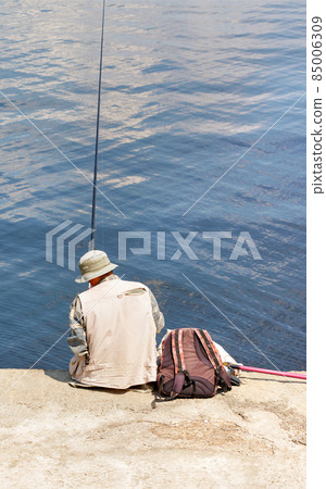 A lone fisherman in a Panama hat sits on a concrete pier with a fishing rod by the water. A lone fisherman in a Panama hat sits on a concrete pier with a fishing rod by the water. 85006309
