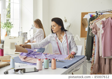 Woman seamstress sit at table design garment in fashion atelier with colleague. Female designer or tailor create clothing piece in workshop, work with fabric textile. Style, dressmaking concept. 85007612