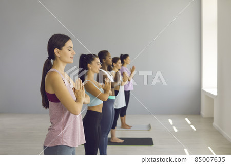 Profile view of different women standing in row making gesture of namaste and meditating with closed eyes restoring energy levels. Concept of exercises that reduce feelings of nervousness and stress. 85007635