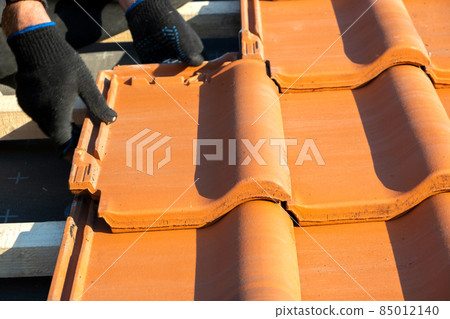 Closeup of worker hands installing yellow ceramic roofing tiles mounted on wooden boards covering residential building roof under construction. 85012140