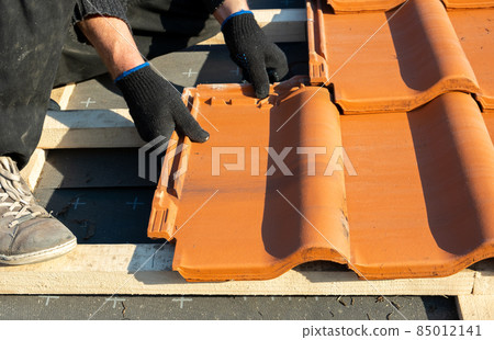 Closeup of worker hands installing yellow ceramic roofing tiles mounted on wooden boards covering residential building roof under construction. 85012141