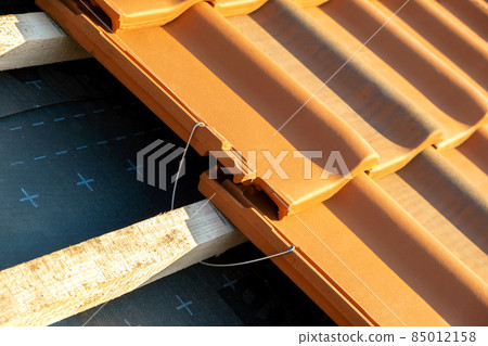 Closeup of metal montage anchor for installation of yellow ceramic roofing tiles mounted on wooden boards covering residential building roof under construction. 85012158