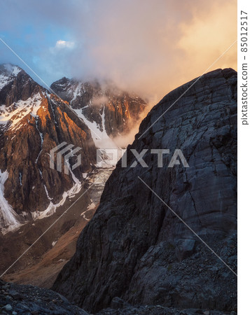 Sunlight in the mountains. Big glacier on top in orange light. Scenic mountain landscape with great snowy mountain range lit by dawn sun among low clouds. Sunlight in the mountains. Big glacier on top in orange light. Scenic mountain landscape with great snowy mountain range lit by dawn sun among low clouds. 85012517
