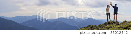 Back view of young tourist couple, athletic man and slim girl standing with raised arms holding hands on rocky mountain top enjoying fantastic panorama. Tourism, traveling and climbing concept. Back view of young tourist couple, athletic man and slim girl standing with raised arms holding hands on rocky mountain top enjoying fantastic panorama. Tourism, traveling and climbing concept. 85012542