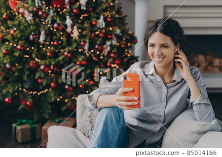 Happy hispanic woman in wireless earphones holding phone, sitting near Christmas tree at home 85014166