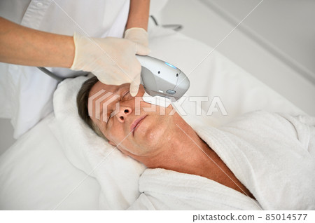 Top view of mature Caucasian man getting masculine skin care at wellness spa centre while beautician doing face lifting anti-aging beauty treatment on his face using ultrasonic medical apparatus Top view of mature Caucasian man getting masculine skin care at wellness spa centre while beautician doing face lifting anti-aging beauty treatment on his face using ultrasonic medical apparatus 85014577