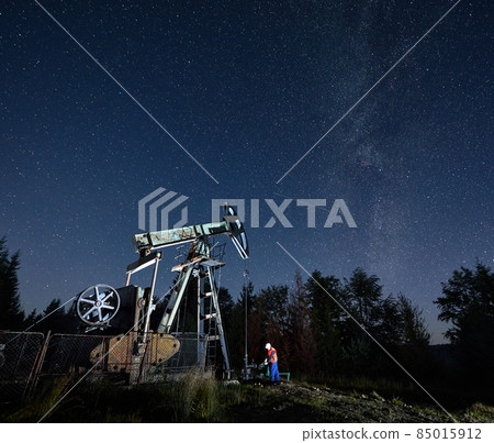 Mechanic working in pump borehole in the evening. Pumping oil and gas from subsoil of ground. Petroleum rig station located in the middle of forest under nightly sky fulled by stars and Milky way. 85015912