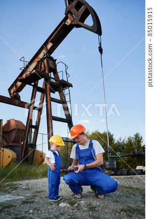Engineer father introducing his son to work tool. Adult man and preschool child in workwears and helmets together looking to adjustable wrench on background of pump for oil production. Engineer father introducing his son to work tool. Adult man and preschool child in workwears and helmets together looking to adjustable wrench on background of pump for oil production. 85015915