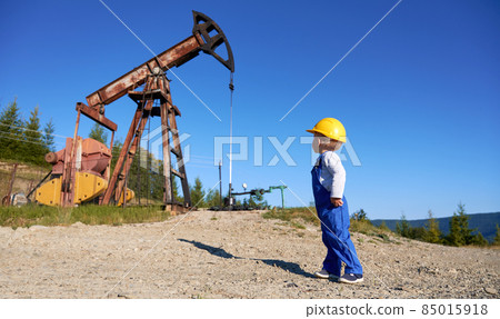 Young boy in blue coverall and yellow casque looking at offshore machine in nature environment and thinking about technician education, male learning system of drilling rig and mining pump Young boy in blue coverall and yellow casque looking at offshore machine in nature environment and thinking about technician education, male learning system of drilling rig and mining pump 85015918