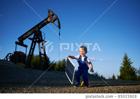 Young kid engineer having fun outdoors in field with oil pump jack at sunny summer day. Smiling child holding small solar panel. Alternative fotovoltaic ecoenergy conception. Young kid engineer having fun outdoors in field with oil pump jack at sunny summer day. Smiling child holding small solar panel. Alternative fotovoltaic ecoenergy conception. 85015928