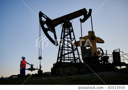 Oilman released excess pressure of gas into oil pipeline and watching as oil pump working. Worker ensuring safe extraction of oil. Oil drilling derrick at oilfield for petroleum production from ground Oilman released excess pressure of gas into oil pipeline and watching as oil pump working. Worker ensuring safe extraction of oil. Oil drilling derrick at oilfield for petroleum production from ground 85015930