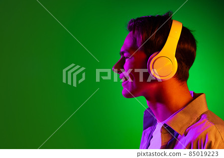 Portrait of young man, student listening to music in headphones isolated on green studio background in neon light 85019223