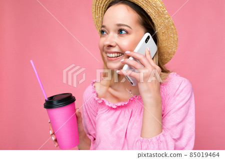 Happy young woman talking on mobile phone while having coffee on pink background Happy young woman talking on mobile phone while having coffee on pink background 85019464