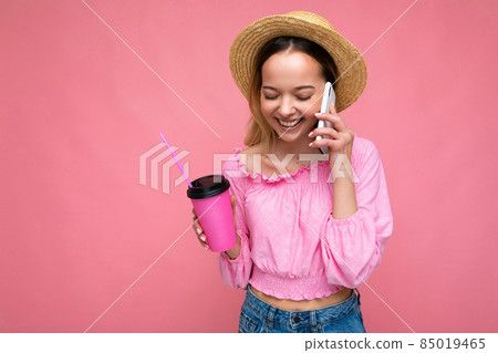 Happy young woman talking on mobile phone while having coffee on pink background Happy young woman talking on mobile phone while having coffee on pink background 85019465
