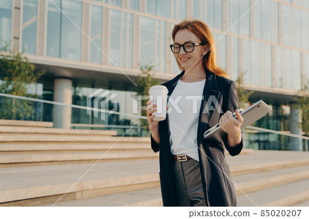 Stylish businesswoman with laptop drinking coffee from disposable paper cup while going at work 85020207