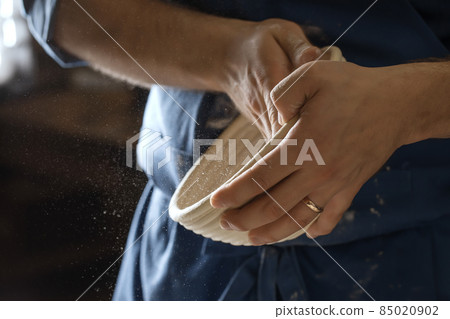 Male baker lightly flouring proofing basket for home made sourdough bread. 85020902