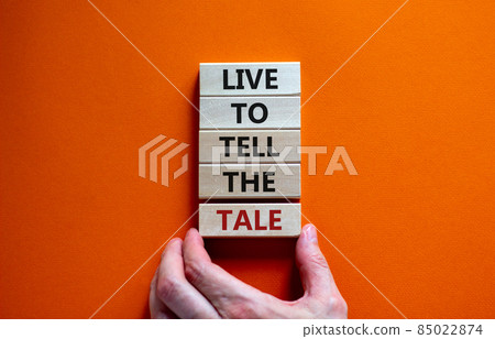 Live to tell the tale symbol. Wooden blocks with words 'live to tell the tale' on beautiful orange background, copy space. Businessman hand. Business and live to tell the tale concept. Live to tell the tale symbol. Wooden blocks with words 'live to tell the tale' on beautiful orange background, copy space. Businessman hand. Business and live to tell the tale concept. 85022874