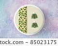 Beautiful and delicious white designer Christmas cake on the table with winter frosty pink and blue patterns with beautiful smiling Christmas trees in the form of chocolate cookies - top view 85023175