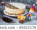 A big beautiful Christmas round mousse cake with festive patterns, tinsel and candles in jars on the table with a snowman in a cap of rainbow colors and a pine cone 85023176