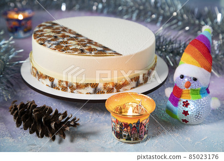 A big beautiful Christmas round mousse cake with festive patterns, tinsel and candles in jars on the table with a snowman in a cap of rainbow colors and a pine cone A big beautiful Christmas round mousse cake with festive patterns, tinsel and candles in jars on the table with a snowman in a cap of rainbow colors and a pine cone 85023176