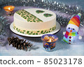 New Year's snowman in a scarf and a rainbow - colored hat with a Christmas Eve chocolate cake with beautiful Christmas tree cookies and Christmas decor - candles, pine cones, tinsel 85023178
