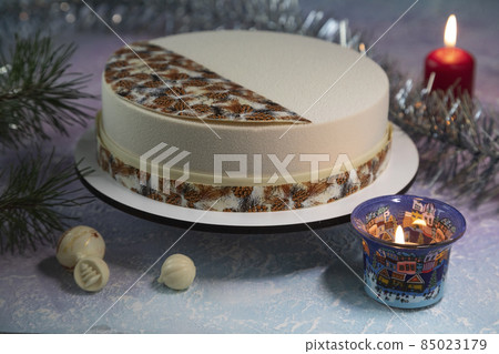 Christmas Eve - a beautiful cake on the table with a pine cone, candle lights, a garland of tinsel and chocolate toys for decorating a mousse cake - a concept for a Christmas card Christmas Eve - a beautiful cake on the table with a pine cone, candle lights, a garland of tinsel and chocolate toys for decorating a mousse cake - a concept for a Christmas card 85023179