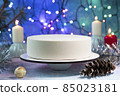 Christmas cake made of French mousse surrounded by garlands, burning candles, a ball on the Christmas tree, pine branches and cones and tinsel on a frosty blue background 85023181