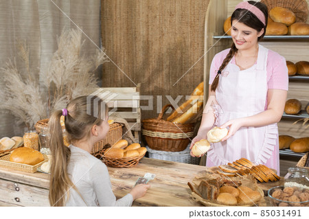 A little girl came to buy rolls at the bakery store. An attractive saleswoman serves the bread behind the counter. Family bakery shop. A little girl came to buy rolls at the bakery store. An attractive saleswoman serves the bread behind the counter. Family bakery shop. 85031491