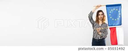 Young girl with the flag of the European Union and Poland. The student is holding two flags in her hand. Woman with flags on white background. Panoramic frame. Young girl with the flag of the European Union and Poland. The student is holding two flags in her hand. Woman with flags on white background. Panoramic frame. 85031492
