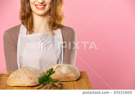 An attractive young saleswoman holds two plump breads decorated with herbs and ears of ripe grain on a wooden board. Seamless light pink background. 85031609