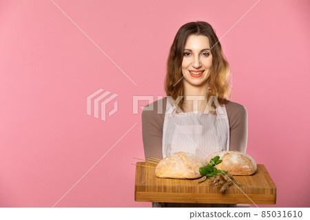 An attractive young saleswoman holds two plump breads decorated with herbs and ears of ripe grain on a wooden board. Seamless light pink background. 85031610