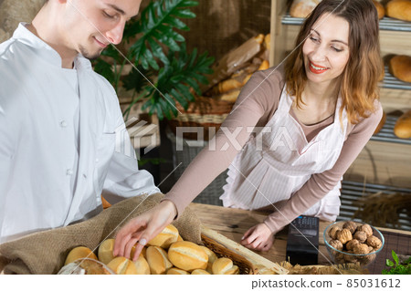 As fresh bread is delivered by a handsome baker, the attractive saleswoman offers one bun. Family bakery shop with multi-generation traditions. 85031612