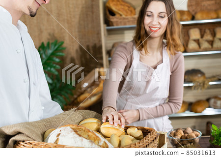 As fresh bread is delivered by a handsome baker, the attractive saleswoman offers one bun. Family bakery shop with multi-generation traditions. 85031613