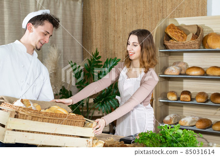 A handsome baker brings a whole box of fresh rolls to the bakery shop and an attractive saleswoman takes all the goods from him. Family bakery shop with multi-generation traditions. 85031614