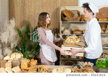 A handsome baker brings a whole box of fresh rolls to the bakery shop and an attractive saleswoman takes all the goods from him. Family bakery shop with multi-generation traditions. 85031624