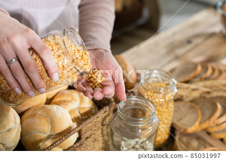 A young attractive saleswoman pours dried corn kernels from a glass container on her hand. Family bakery shop with multi-generation traditions. A young attractive saleswoman pours dried corn kernels from a glass container on her hand. Family bakery shop with multi-generation traditions. 85031997