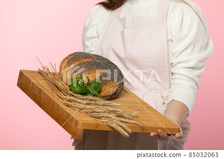 A young attractive saleswoman from a bakery shop holds a round loaf of bread sprinkled with poppy seeds and a pinch of fresh green herb leaves and rye ears on a wooden tray. The whole on a uniform 85032063