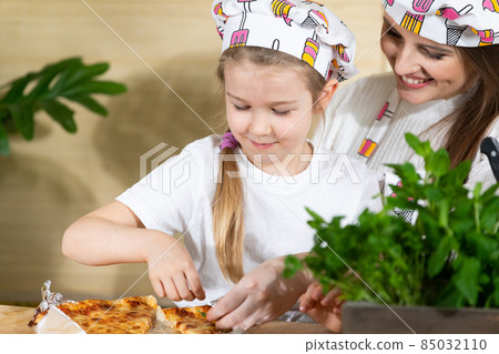 Decorating by mother and her five-year-old daughter freshly baked pizza with green leaves of various herbs. Mom and daughter moments together in the home kitchen. 85032110
